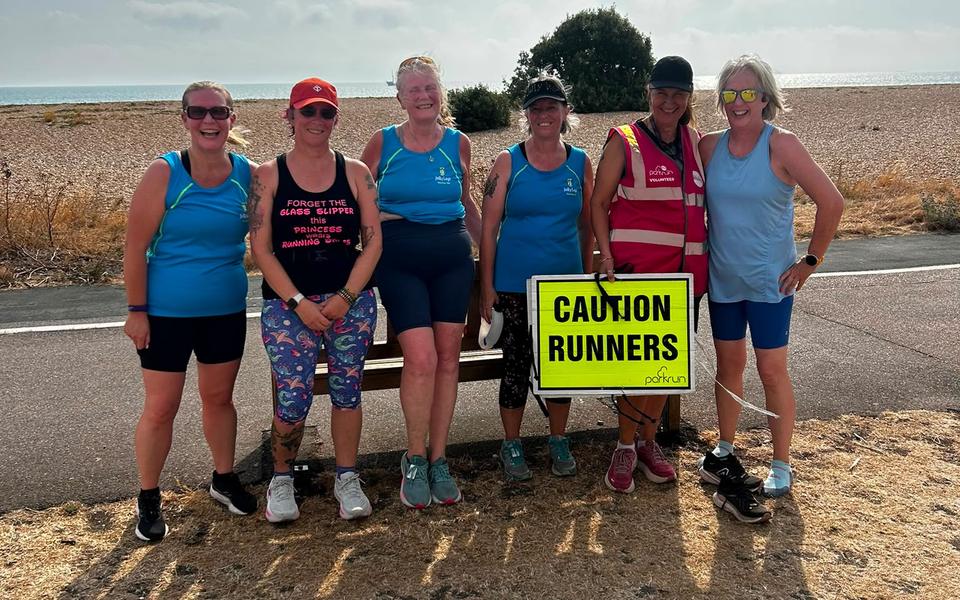 Six smiling Jelly Legs members on the seafront at parkrun, one in a pink volunteer vest holding a Caution Runners sign